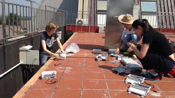 Three students sit on a tiled rooftop working on electronics