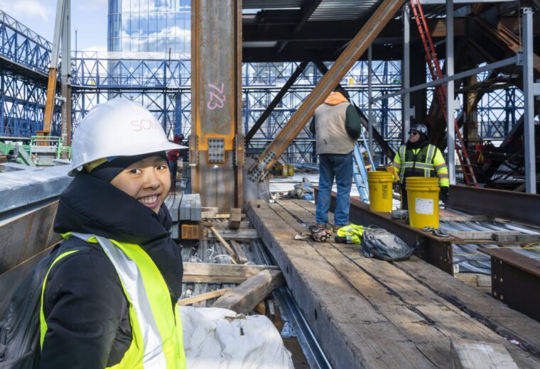 Woman in construction hat and vest at a construction site. She is looking back over her shoulder at the camera and smiling.
